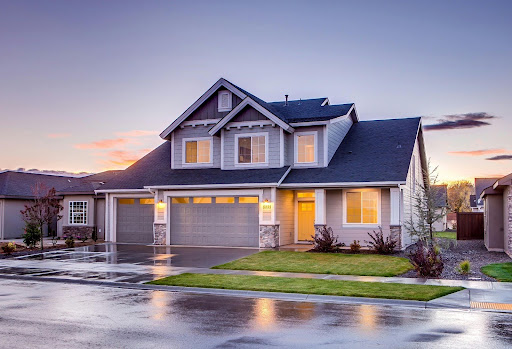 Two-story suburban house with a three-car garage, gray siding, and lit windows—recently updated through home renovation—photographed at dusk after rain with a wet driveway and neatly trimmed lawn.