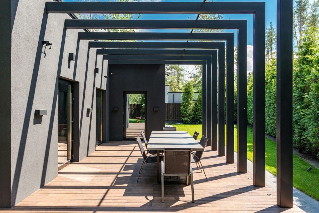 Modern outdoor patio designed for stylish outdoor living spaces, featuring a dining table and chairs beneath rectangular metal pergola beams attached to a dark house, all overlooking a lush green lawn.