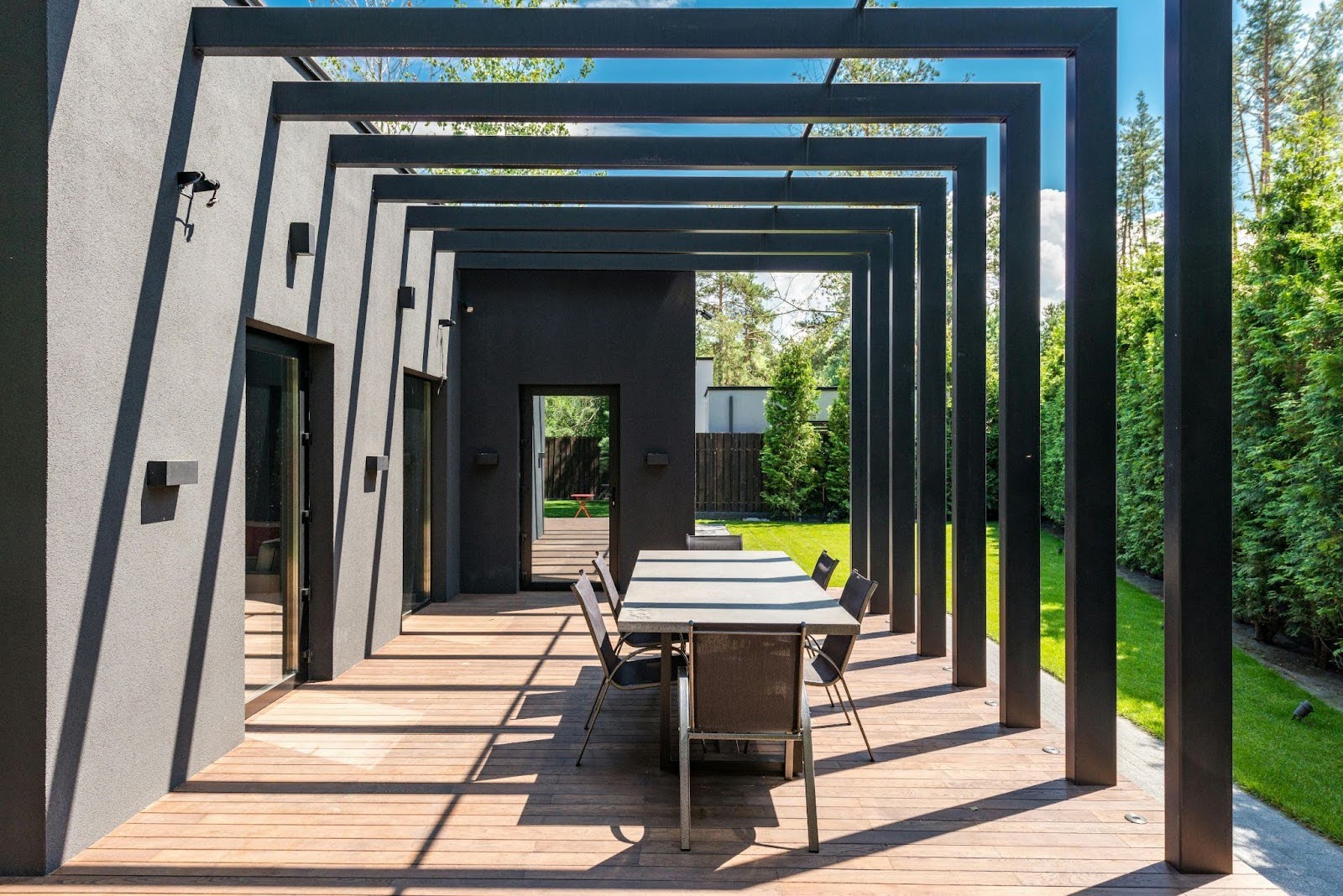 Modern outdoor patio designed for stylish outdoor living spaces, featuring a dining table and chairs beneath rectangular metal pergola beams attached to a dark house, all overlooking a lush green lawn.