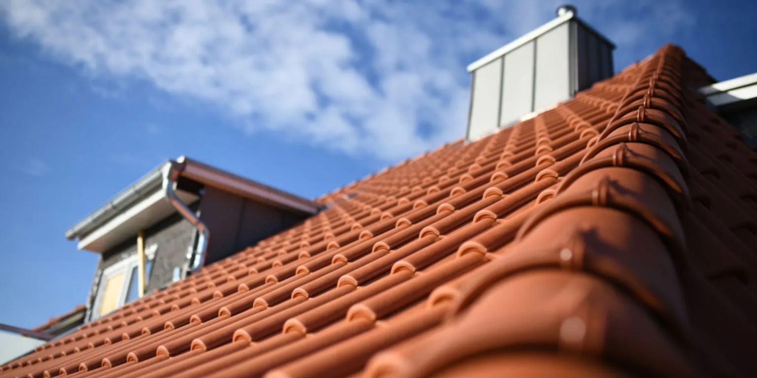 Close-up of a sloped roof with red clay tiles, a dormer window, and a blue sky with scattered clouds in the background.