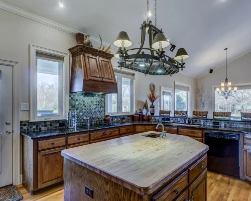 Spacious kitchen with wooden cabinets, a large island with a built-in sink, tiled backsplash, and modern light fixture under vaulted ceiling with large windows.