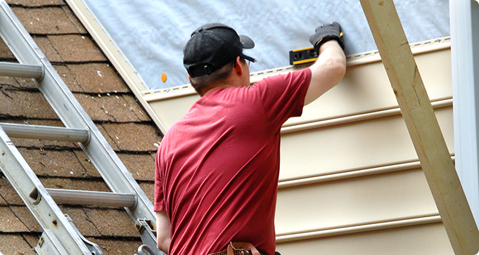 A person in a red shirt and cap is working on siding installation on a house roof. A ladder is positioned against the roof.