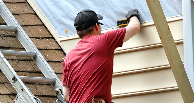 A person wearing a red shirt and black cap installs siding on a house using a tool, with a ladder leaned against the roof nearby.