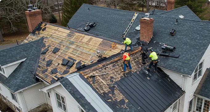 Three workers in safety gear replace shingles on a house roof; part of the roof is stripped down to the wooden decking while the rest has new shingles installed.