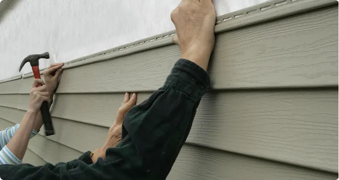 Two people are installing horizontal vinyl siding on an exterior wall; one is holding siding in place while the other uses a hammer to secure it.