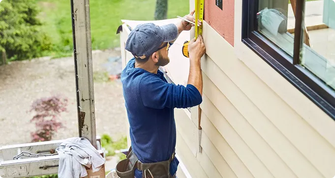 A man in a blue shirt and cap uses a tape measure to measure siding on the exterior of a house near a window.
