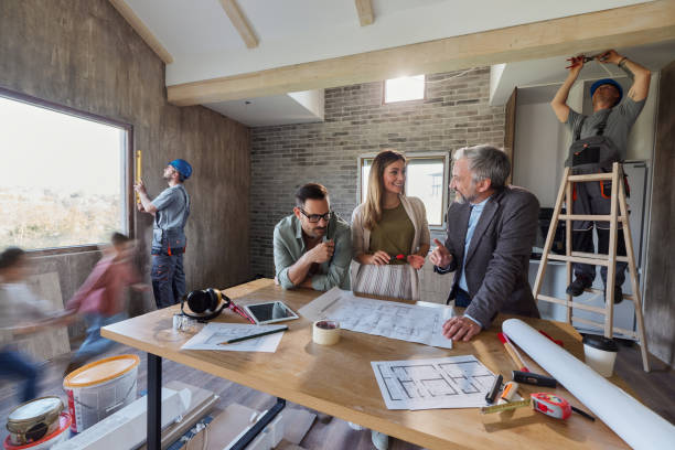 Group discussing plans at a construction site with tools on the table. Two workers measure windows, another installs lighting on a ladder. Blurred individuals walk past.
