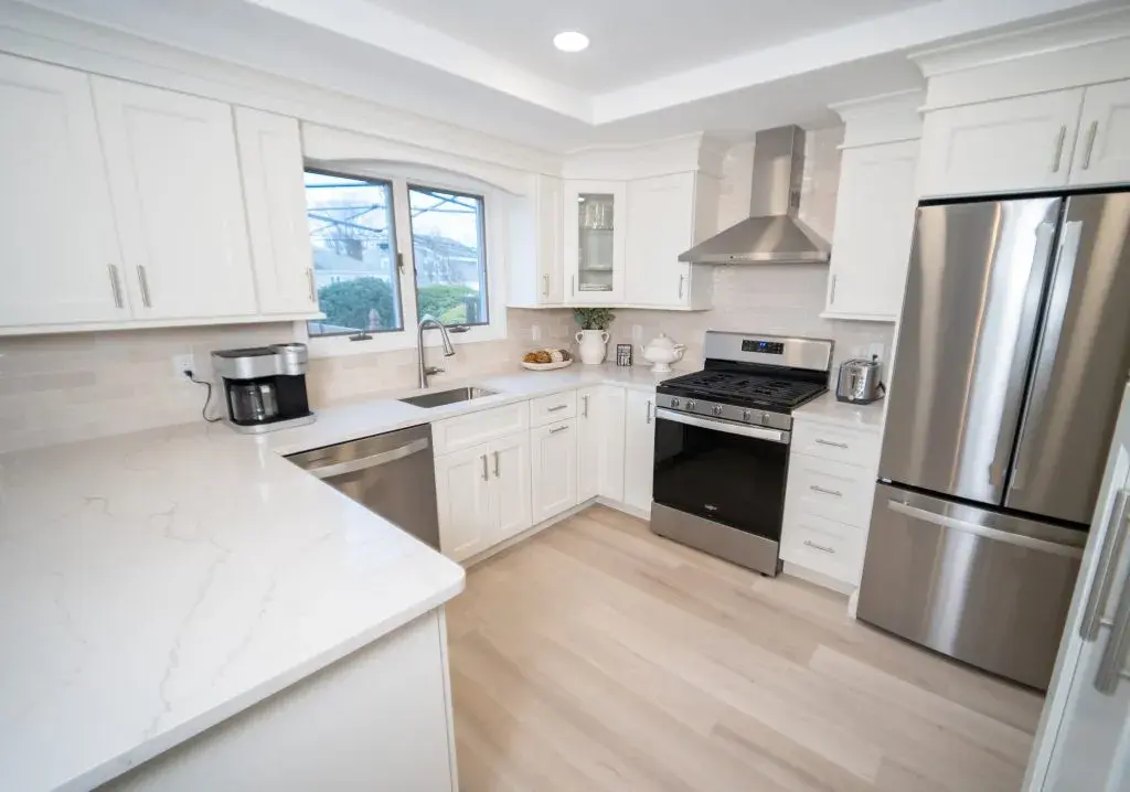Modern kitchen featuring selective remodeling with white cabinets, stainless steel appliances, and light wood flooring. The space offers ample countertop area, a window above the sink for natural light, and a sleek range hood above the stove.