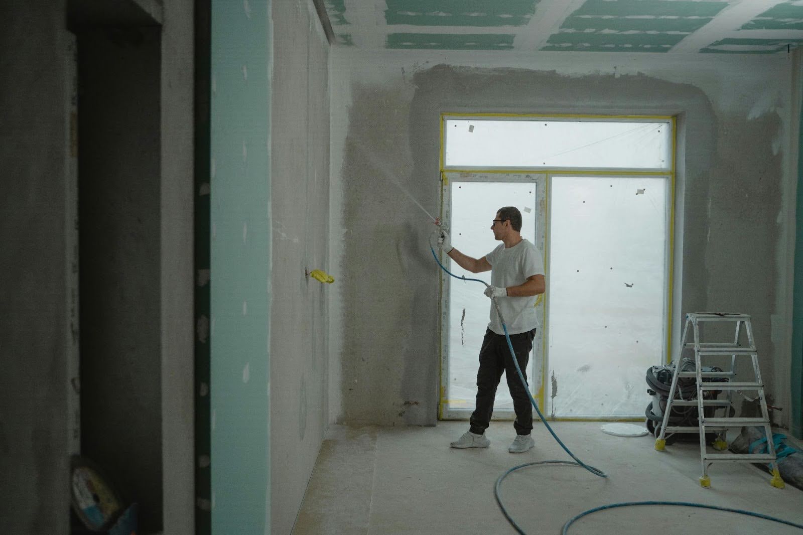 A person, likely one of the home renovation contractors, uses a spray gun to paint or coat the walls of an unfinished room, with a ladder and tools nearby and a large window in the background.