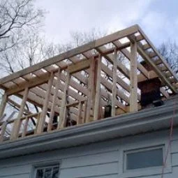 Wooden framing for a house addition or extension is being constructed on top of an existing roof, with exposed beams and rafters visible against the sky.