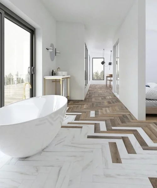 Modern bathroom with a freestanding white tub, herringbone patterned tile flooring in marble and wood tones, a sink, and large windows letting in natural light.