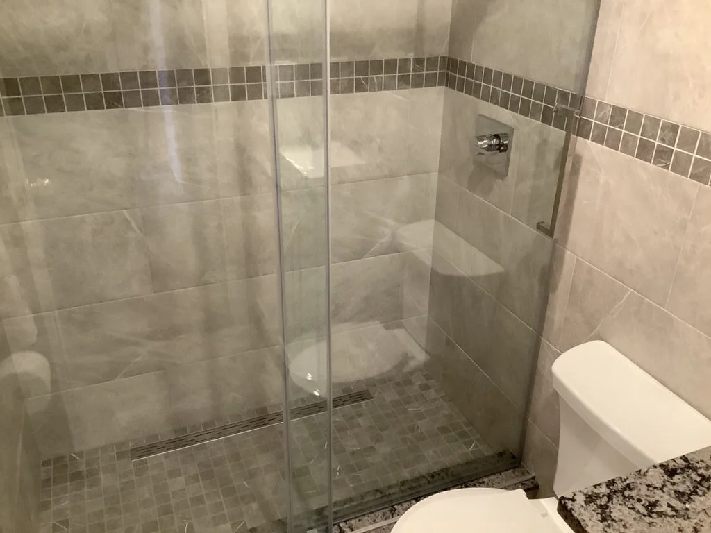 Modern bathroom with a glass-enclosed shower, grey tile walls and floor, built-in shower shelf, and a white toilet in the foreground.