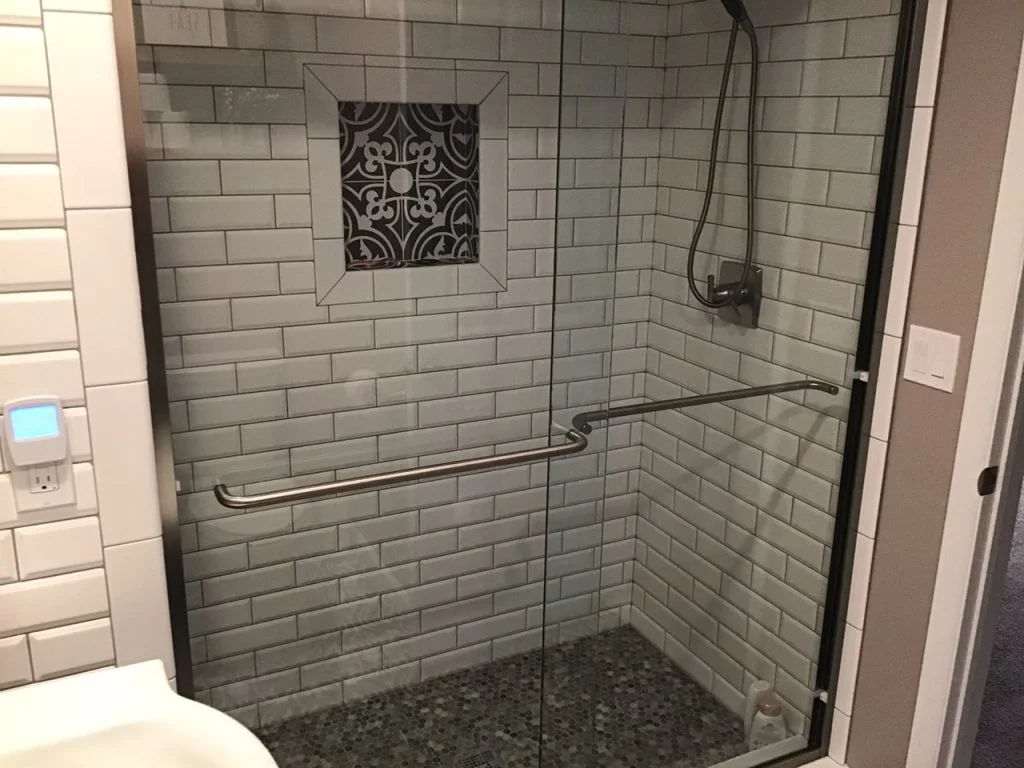 Modern shower with white subway tiles, glass sliding door with metal handle, patterned tile accent, wall-mounted showerhead, and pebble-style floor.