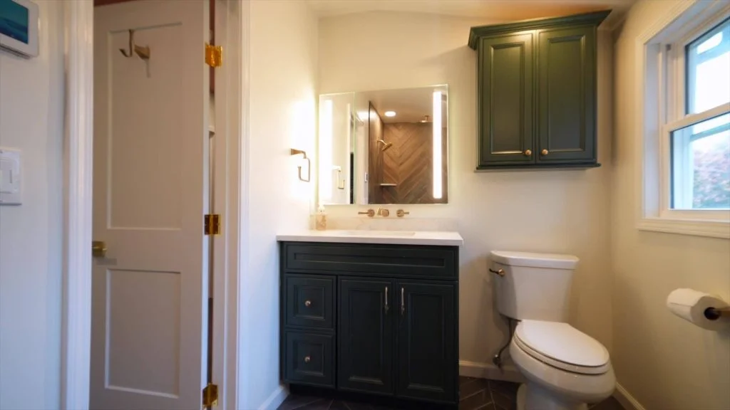 A modern bathroom with a dark green vanity, a wall-mounted cabinet, a toilet, a lighted mirror, and a window. The door is partially open, revealing hooks on the back.