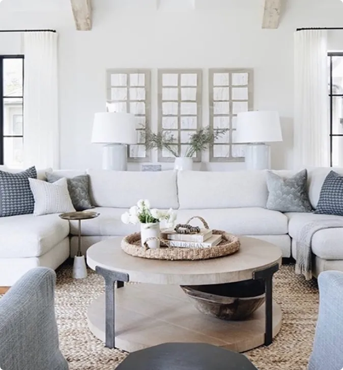 A modern living room with a white sectional sofa, patterned pillows, a round wooden coffee table, two table lamps, and three window-pane wall art pieces.