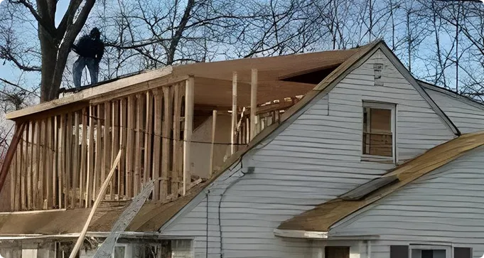 A person stands on a tree branch next to a partially constructed second story on a house, with exposed framing and roofing materials visible.