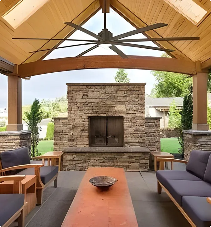 Covered outdoor patio with a stone fireplace, wooden ceiling, large ceiling fan, modern wooden furniture, and a long coffee table. Green lawn and trees visible in the background.