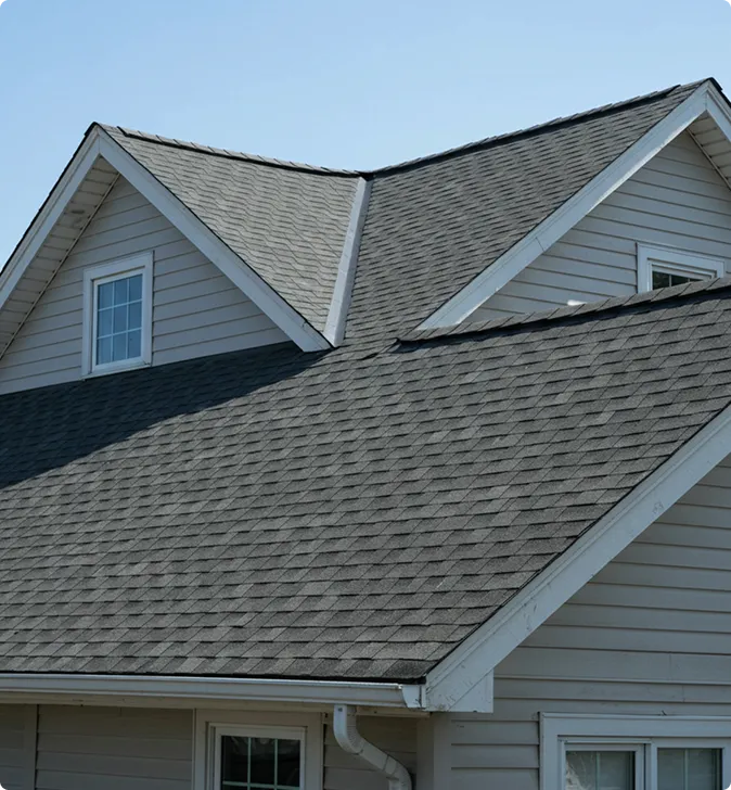 A house with grey asphalt shingle roofing, multiple dormer windows, light-colored siding, and visible white gutters under a clear sky.