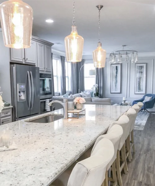 Bright modern kitchen with a large white granite island, white barstools, pendant lights, stainless steel appliances, and an open view to a living room.