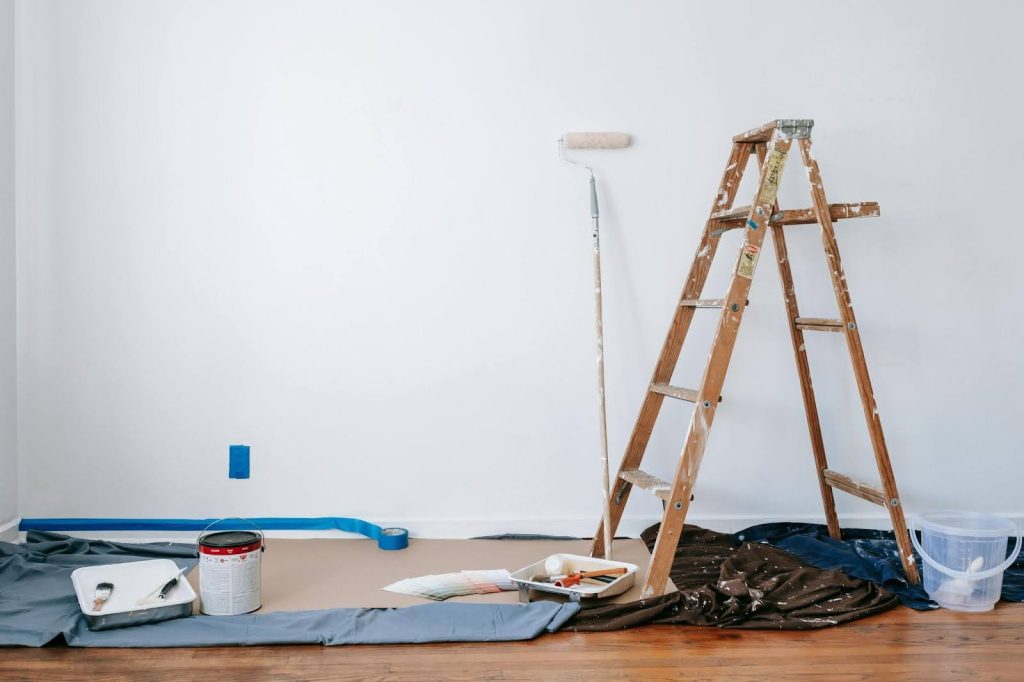 A wooden ladder, paint supplies, and protective coverings are arranged in front of a white wall being prepared for painting.