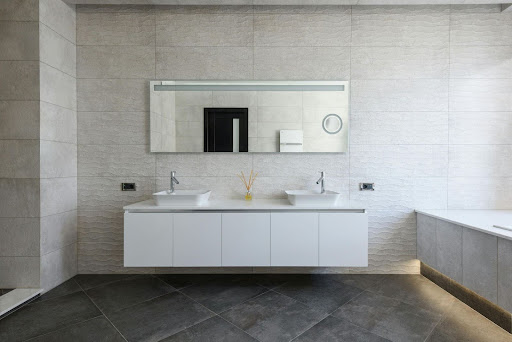 Modern bathroom with large mirror, double sinks on a floating vanity, neutral tiled walls and floors, and a bathtub on the right.