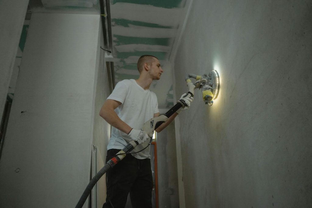 A person wearing gloves and a white shirt uses a wall sander to smooth a surface in an unfinished room.