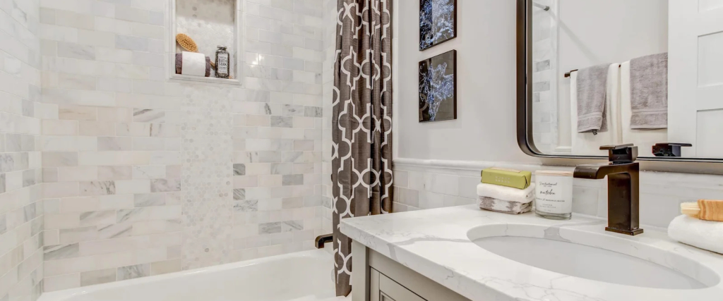 Modern bathroom with a marble countertop, under-mount sink, patterned shower curtain, tiled walls, and a built-in shower shelf holding a brush and bottles.