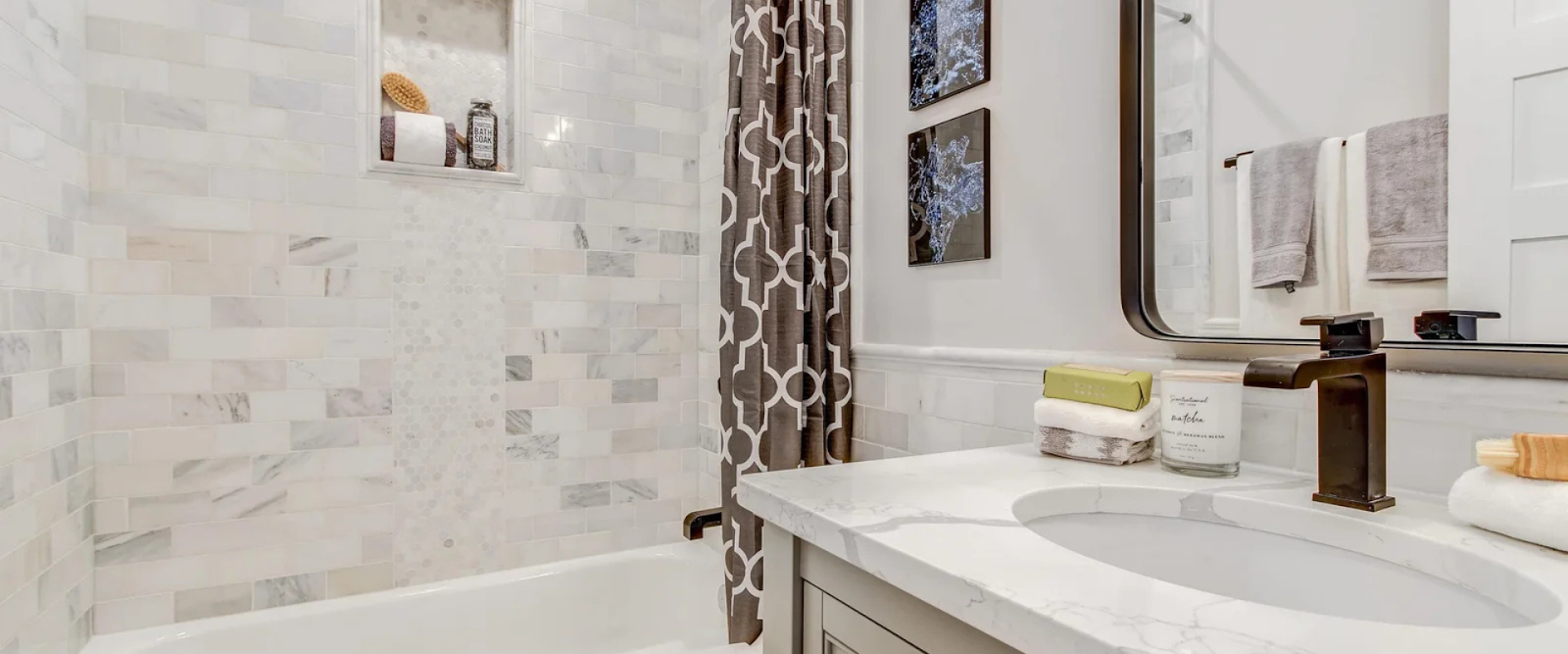 Modern bathroom with a marble countertop, under-mount sink, patterned shower curtain, tiled walls, and a built-in shower shelf holding a brush and bottles.