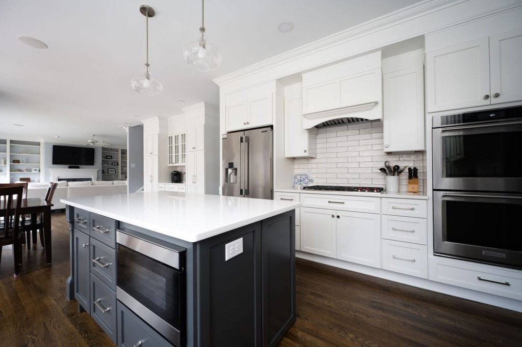 Modern kitchen with white cabinets, stainless steel appliances, a dark island with built-in oven, pendant lights, and hardwood floors, opening to a dining and living area.