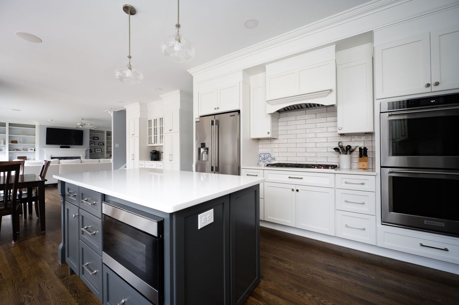 Modern kitchen with white cabinets, stainless steel appliances, a dark island with built-in oven, pendant lights, and hardwood floors, opening to a dining and living area.