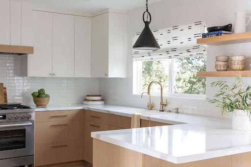 Modern kitchen with light wood and white cabinetry, white countertops, a stainless steel oven, gold faucet, pendant light, and window with patterned shade.