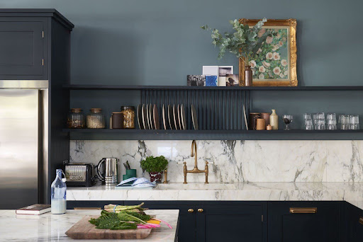 Modern kitchen with dark cabinets, marble backsplash, open shelves holding dishes and glassware, a gold faucet, and a countertop with fresh vegetables and a bottle of milk.