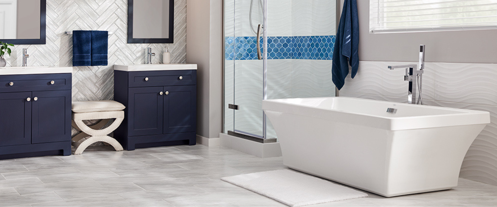 Modern bathroom with a freestanding bathtub, glass shower, double sinks with navy blue cabinets, and light-colored tile flooring.