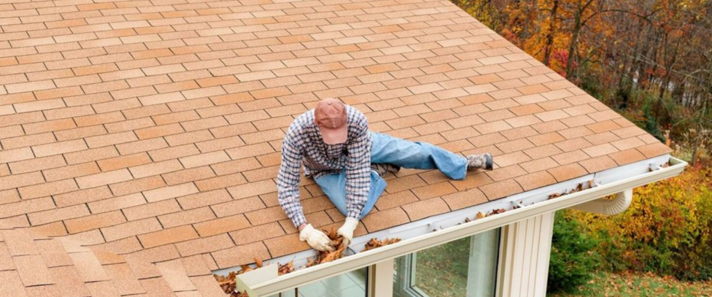 A person in a plaid shirt and cap removes leaves from a houses rain gutter while sitting on a shingled roof.
