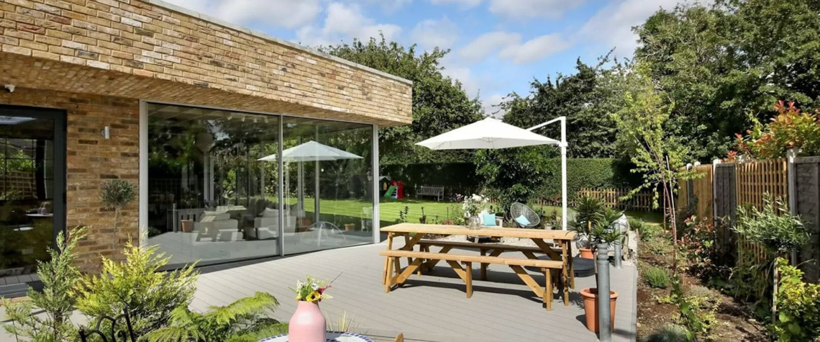 A modern brick house with large glass doors opens onto a patio with a wooden picnic table, white umbrella, and garden plants, overlooking a grassy backyard with trees and a fence.