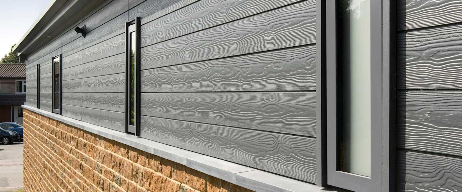 Close-up of a modern building exterior with horizontal gray wood-textured siding above a section of tan brick, featuring black-framed windows.