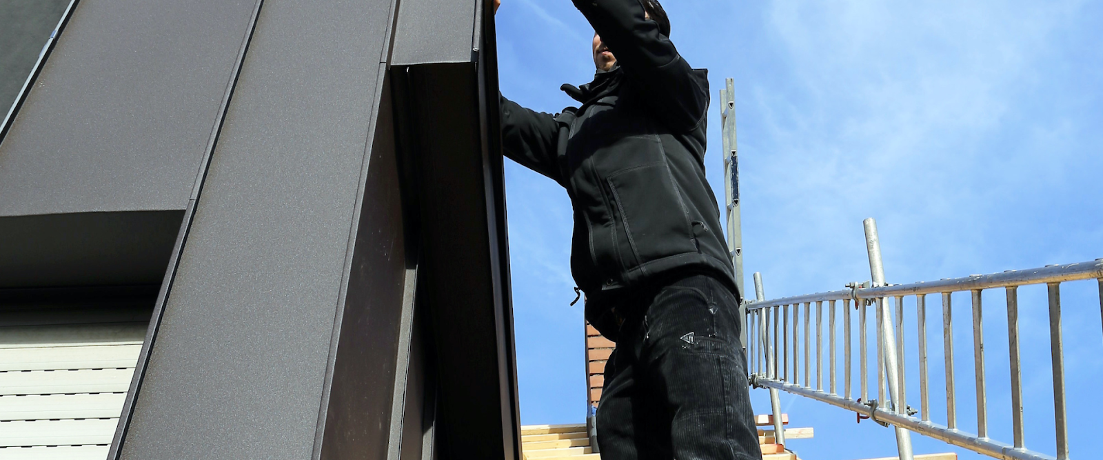A person wearing black work clothes stands on scaffolding next to a modern building, working near the roof on a clear day. A metal ladder is positioned nearby.