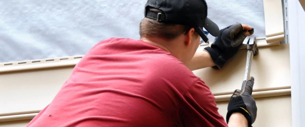 A person wearing gloves, a red shirt, and a black cap installs siding on a building using a hammer.
