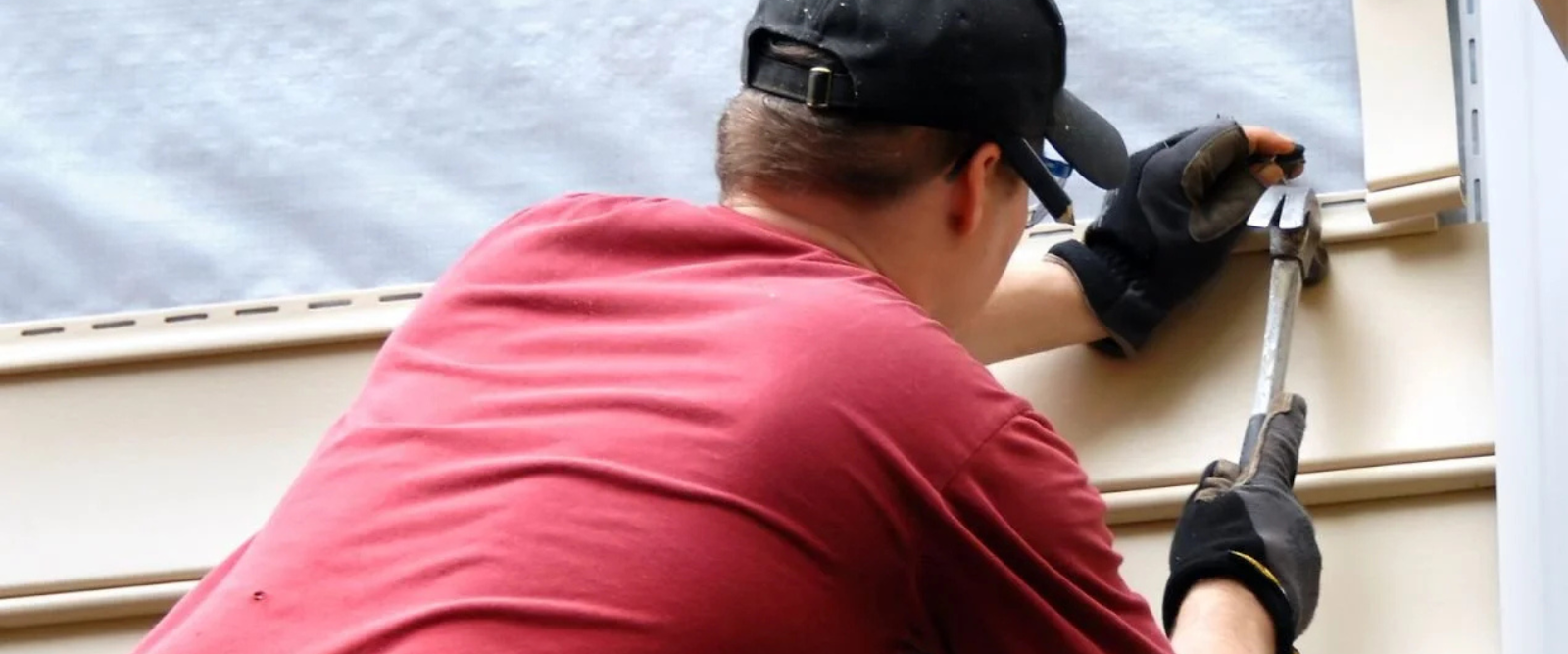 A person wearing gloves, a red shirt, and a black cap installs siding on a building using a hammer.