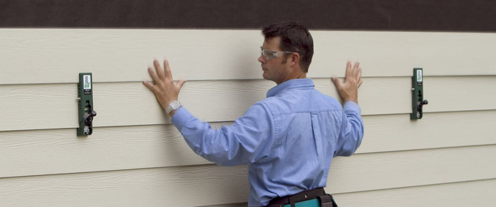 A person wearing safety glasses is installing horizontal siding panels on a building exterior using two green siding tools.