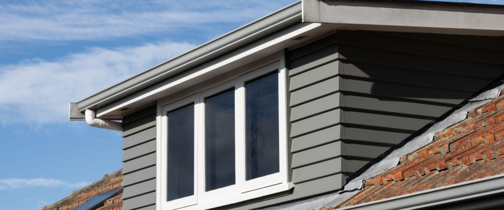 A close-up of a modern dormer window with white framing and grey horizontal cladding on a tiled house roof, under a blue sky with clouds.