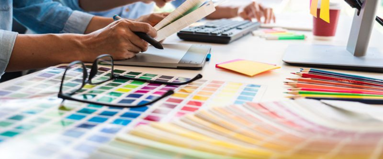Two people work at a desk covered with color swatches, colored pencils, sticky notes, and a keyboard, suggesting a design or creative project.