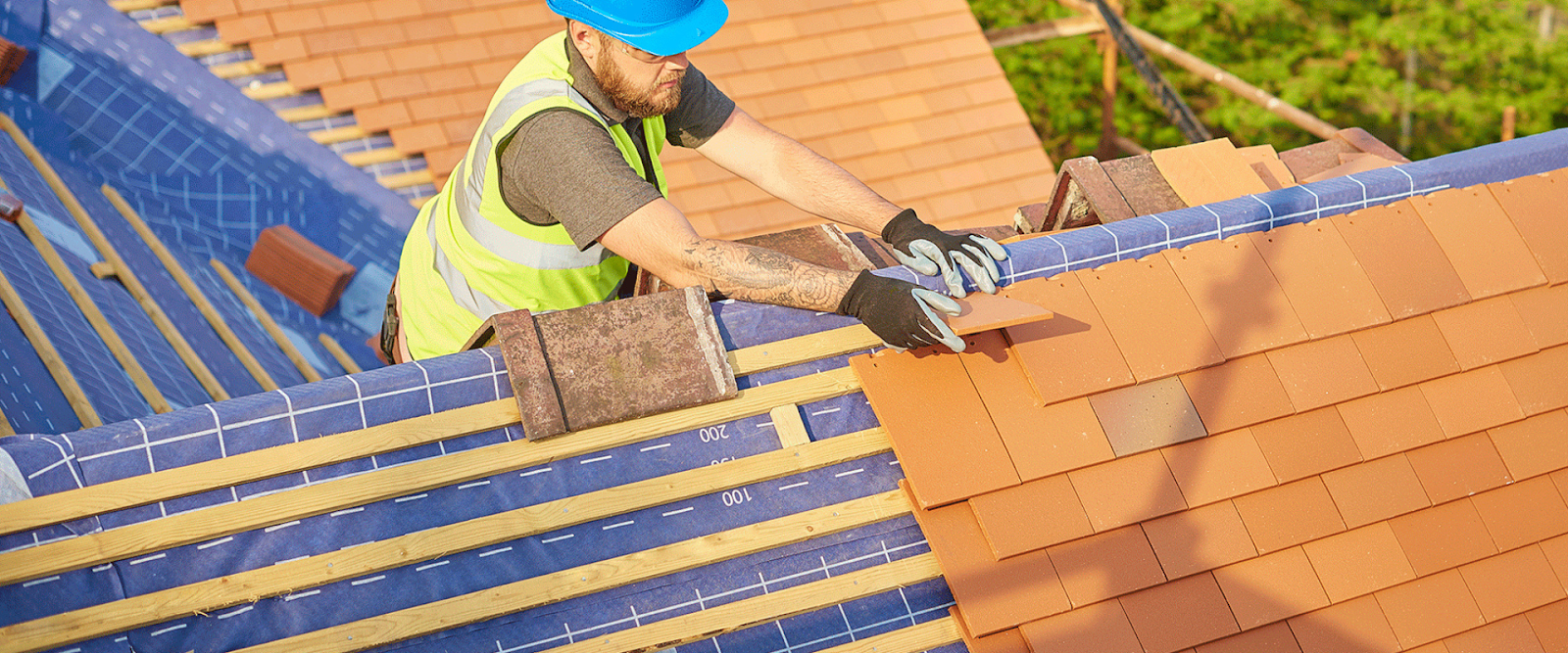 A construction worker wearing a blue helmet and safety vest installs terracotta roof tiles on a wooden framework of a house roof.
