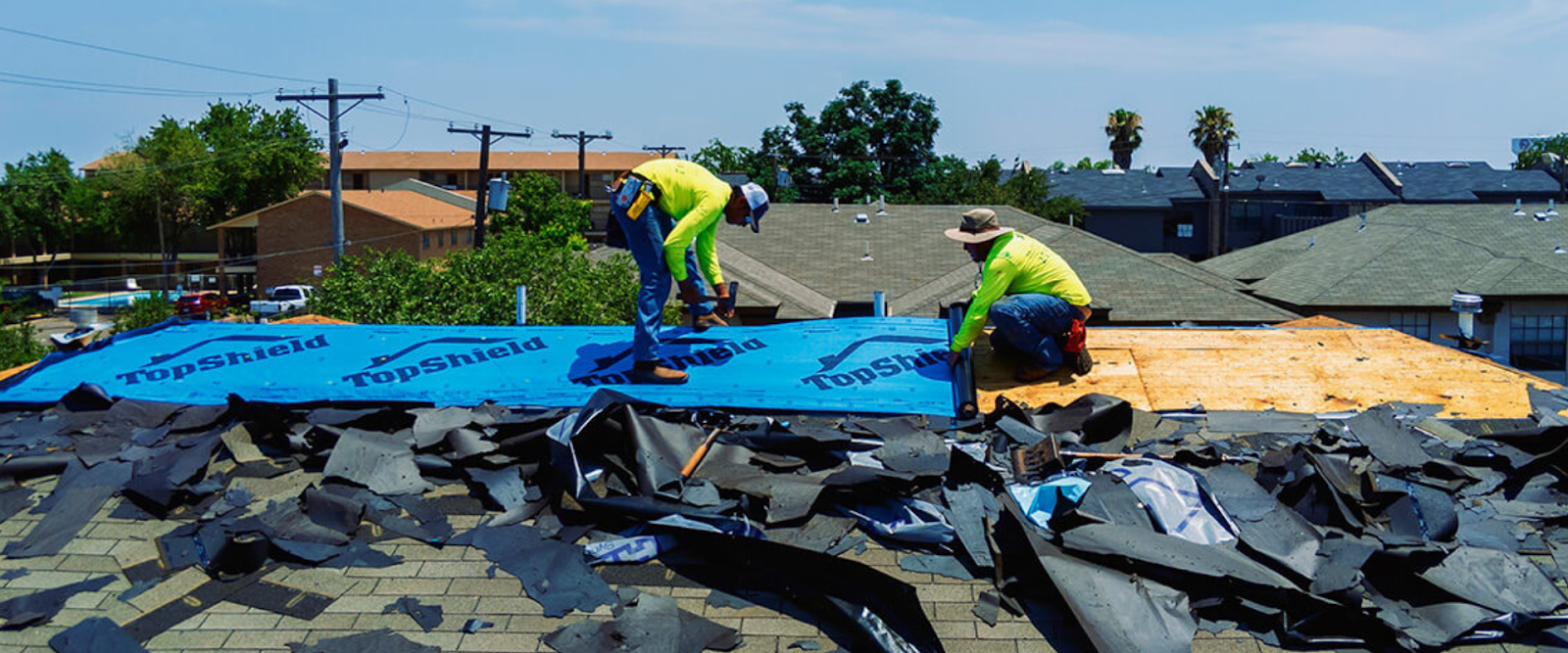 Three workers in yellow shirts repair a roof, removing old shingles and installing new underlayment labeled TopShield on a sunny day. Debris is scattered across the rooftop.