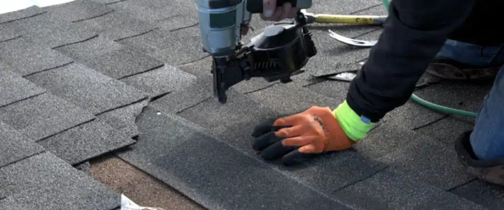 A person wearing gloves installs asphalt shingles on a roof, using a nail gun with tools and other shingles nearby.
