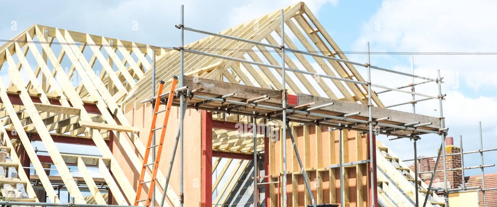 Partially constructed house with exposed wooden framing, scaffolding, and ladders under a blue sky.