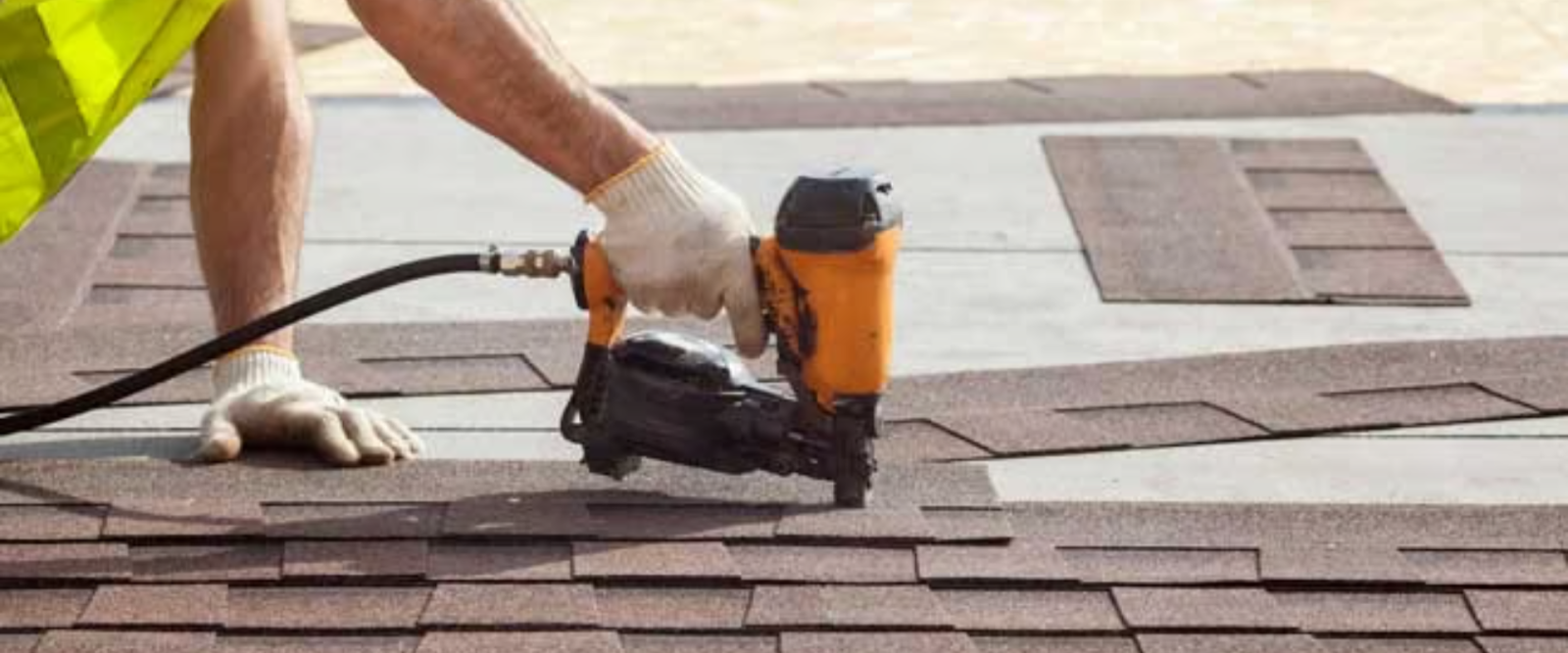 A worker in gloves uses a nail gun to install asphalt shingles on a roof.