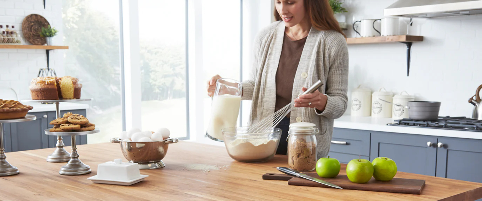 A woman stands in a modern kitchen, pouring milk into a mixing bowl with flour while holding a whisk; baked goods and green apples are on the counter.
