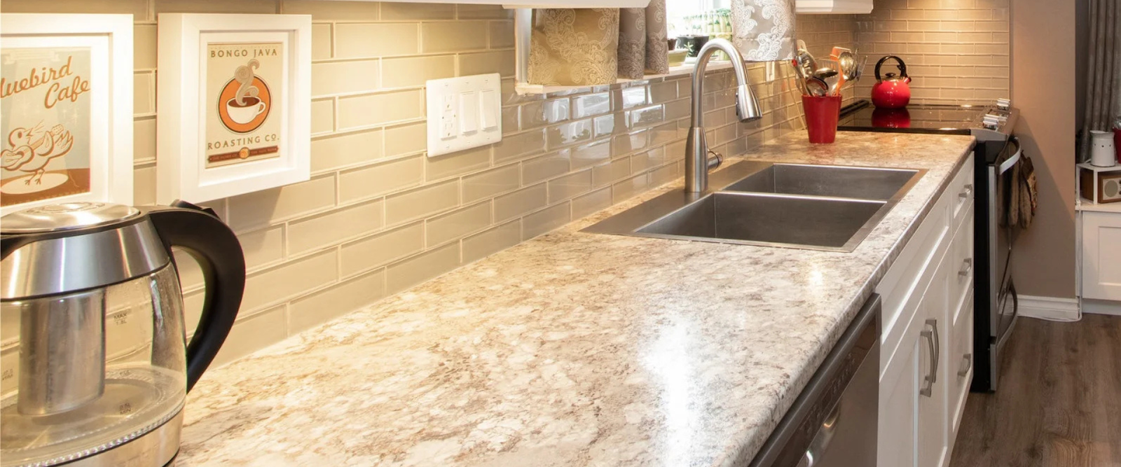 A modern kitchen countertop with a stainless steel sink, electric kettle, red teapot, wall art, and light-colored backsplash tiles.