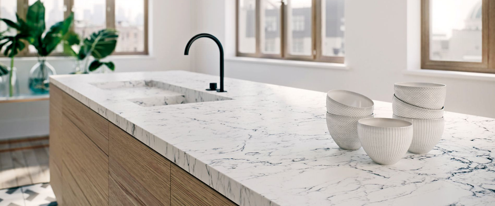 A modern kitchen with a marble countertop, integrated sink, black faucet, and four white textured bowls, with large windows and potted plants in the background.